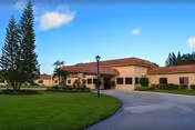 Exterior view of a single-story building with a red-tiled roof, surrounded by green grass, palm trees, and other landscaping under a blue sky with some clouds. A curved driveway leads to the entrance of the building.
