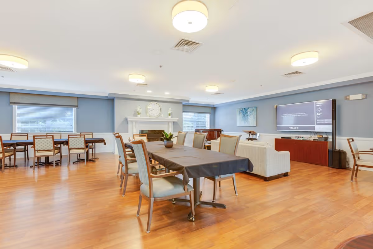 A spacious senior living common area with wooden flooring, multiple tables and chairs, a white fireplace with a clock and decorative items above it, a large flat-screen TV mounted on the wall, and a white couch. The walls are painted light blue with white trim, and there are windows with blinds allowing natural light into the room.