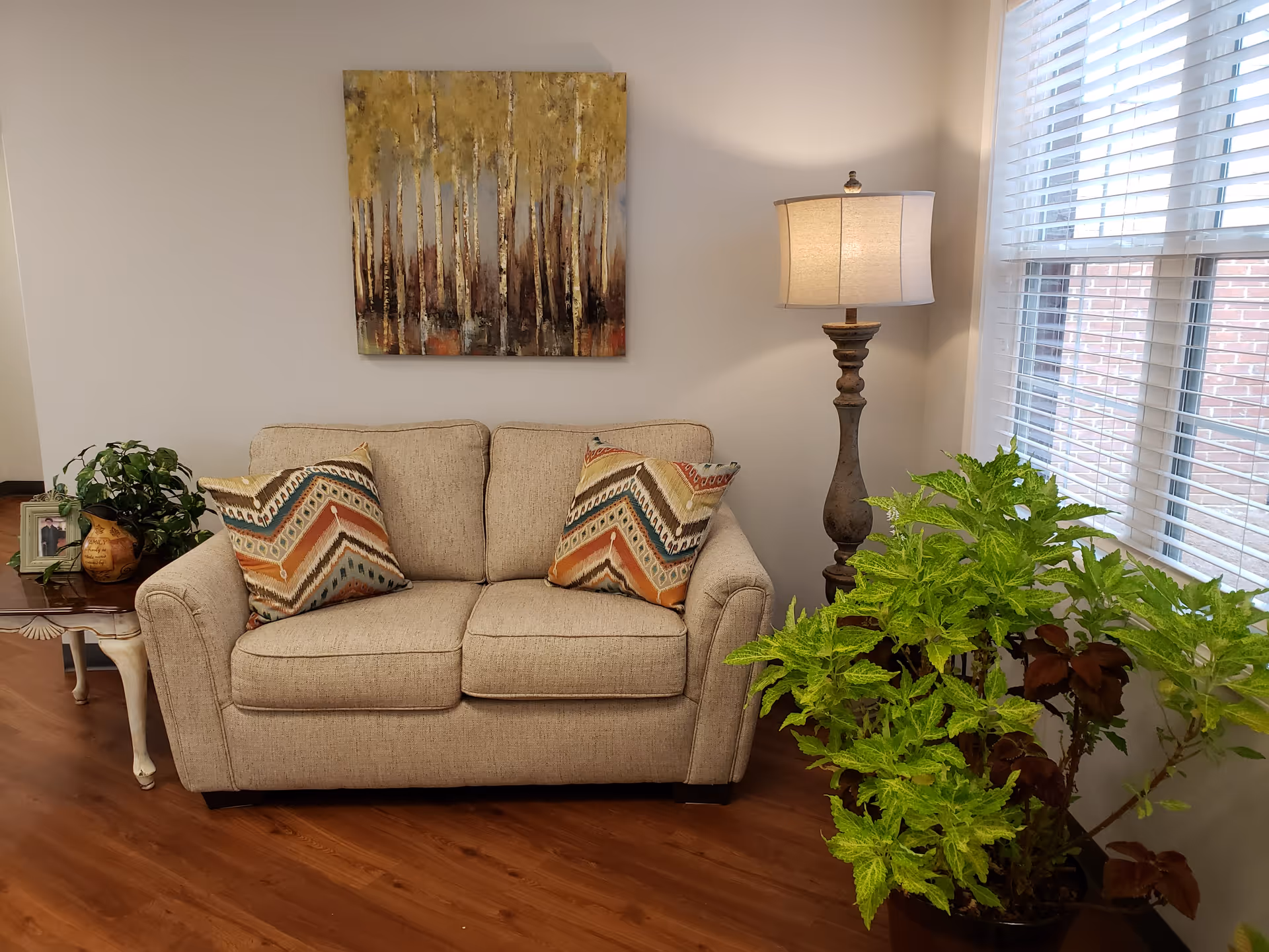 A cozy living room area with a beige loveseat adorned with two patterned throw pillows. Above the loveseat hangs a painting of a forest with tall trees in autumn colors. To the right of the loveseat is a tall floor lamp with a beige shade, and next to it is a large green leafy plant near a window with white blinds. To the left of the loveseat is a small wooden side table with a potted plant and a framed photo. The floor is wooden.