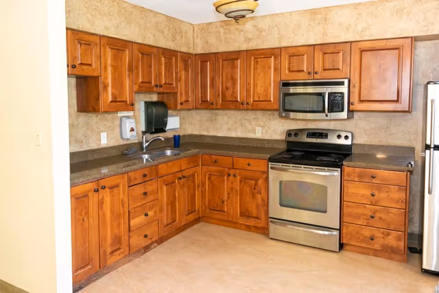 A kitchen with wooden cabinets, a stainless steel oven with a stove, a microwave above the stove, a stainless steel refrigerator, a double sink with a paper towel dispenser mounted on the wall, and a beige countertop and floor.