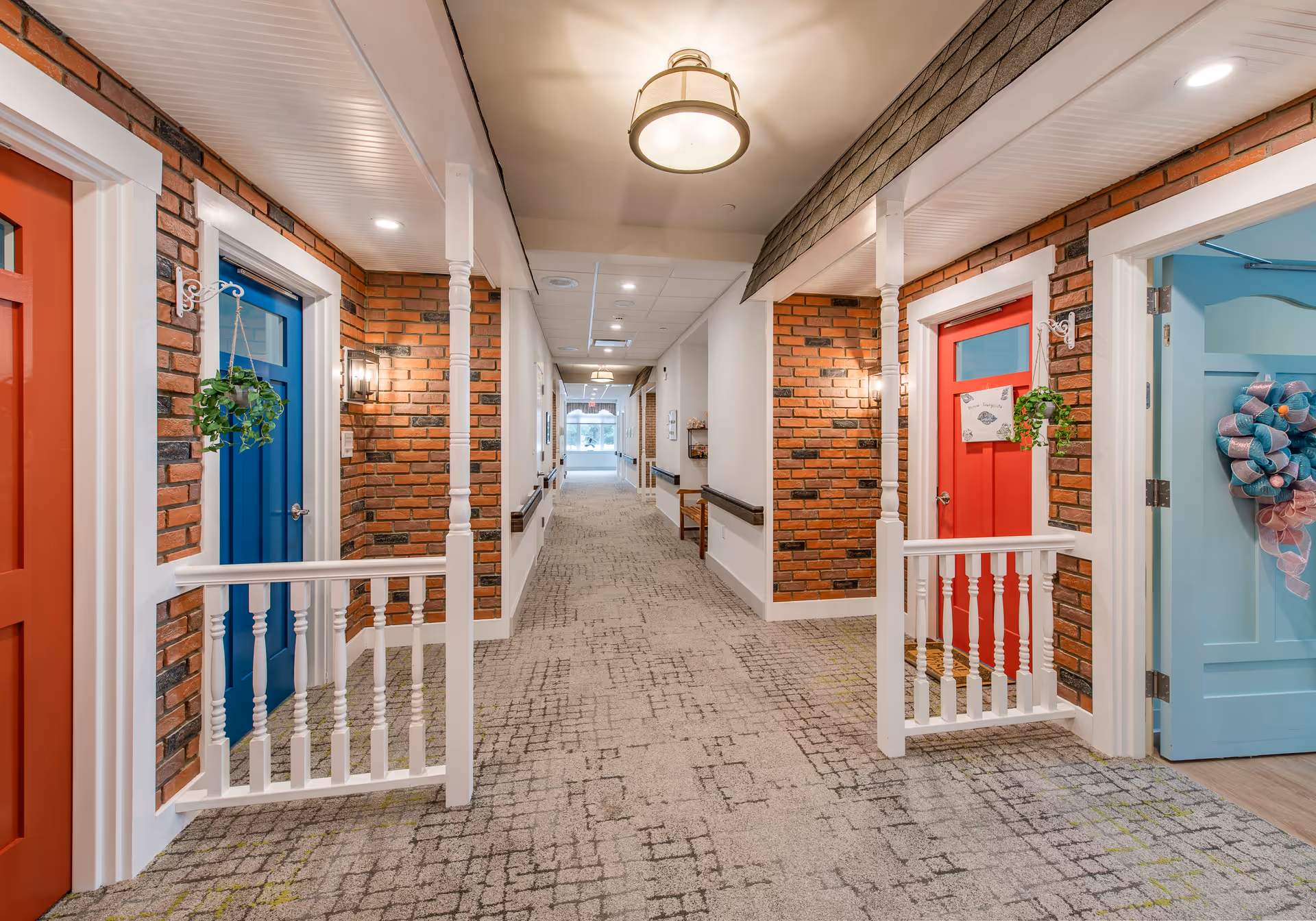 A brightly lit hallway in a senior living facility with brick walls and colorful doors painted red, blue, and light blue. The hallway features small porch-like railings in front of each door, hanging plants, and decorative wreaths. The carpet has a patterned design, and ceiling lights illuminate the space.