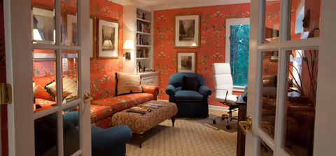 A cozy living room with red patterned wallpaper, a blue armchair, a white office chair, a patterned ottoman, and a red sofa with striped cushions. The room has built-in bookshelves, framed artwork on the walls, and is viewed through glass-paneled double doors.