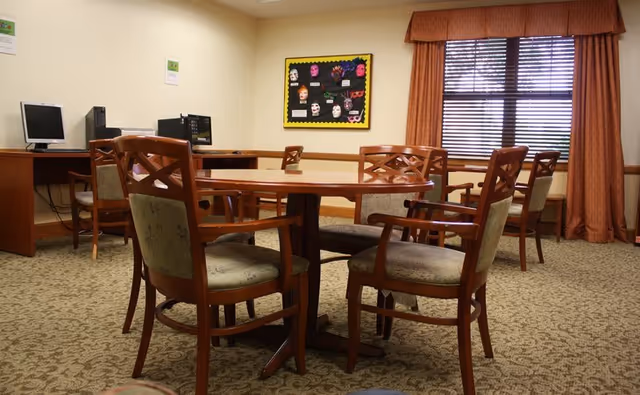 A room with a round wooden table surrounded by six cushioned wooden chairs. In the background, there are two computer desks with desktop computers and chairs. A window with closed blinds and orange curtains is on the right wall. A bulletin board with colorful masks and decorations is mounted on the wall between the window and the computers. The room has beige walls and patterned carpet flooring.