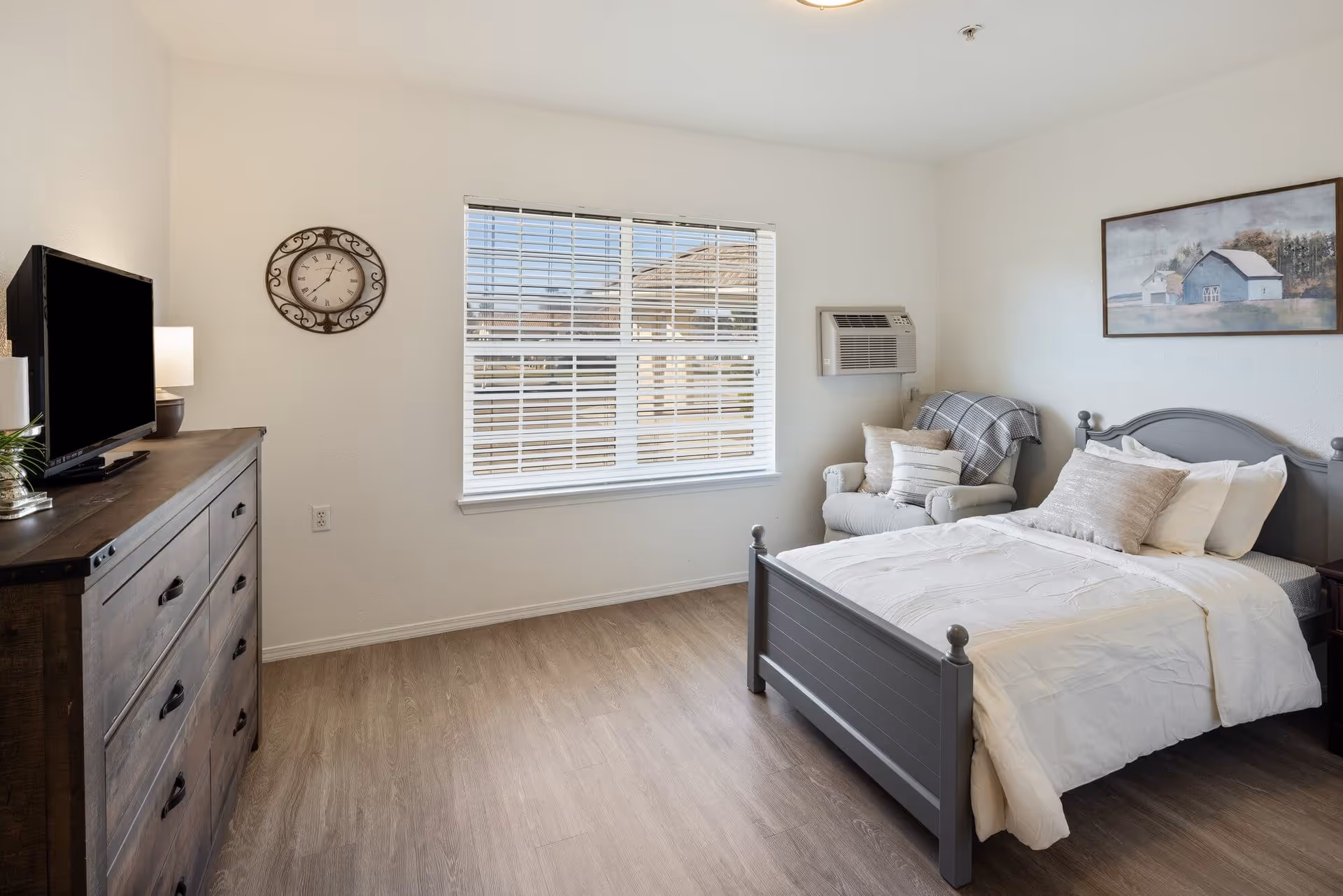 A bright and clean bedroom with a gray wooden bed made with white bedding and multiple pillows. Next to the bed is a cushioned armchair with pillows and a plaid throw blanket. A large window with white blinds lets in natural light. On the left side of the room, there is a wooden dresser with a flat-screen TV on top and a decorative wall clock above it. A framed picture of a barn hangs on the wall above the bed. The room has light wood flooring and white walls.