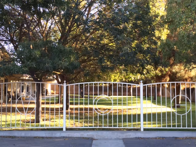White metal fence in front of a grassy yard with large trees and part of a building visible on the left.