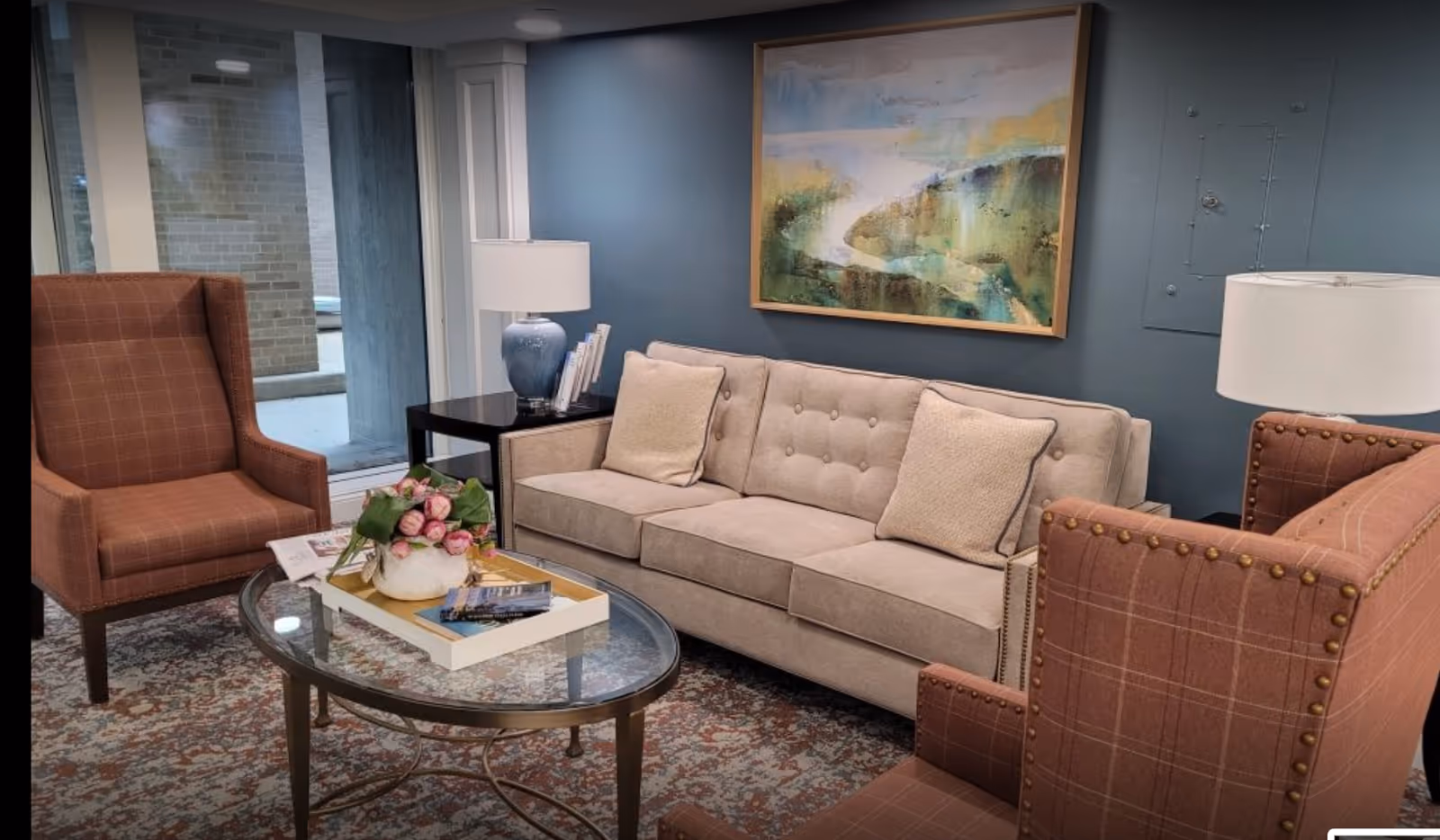 A cozy seating area with a beige tufted sofa, two upholstered armchairs, a glass coffee table holding flowers and books, lamps, and a framed painting on a blue wall.