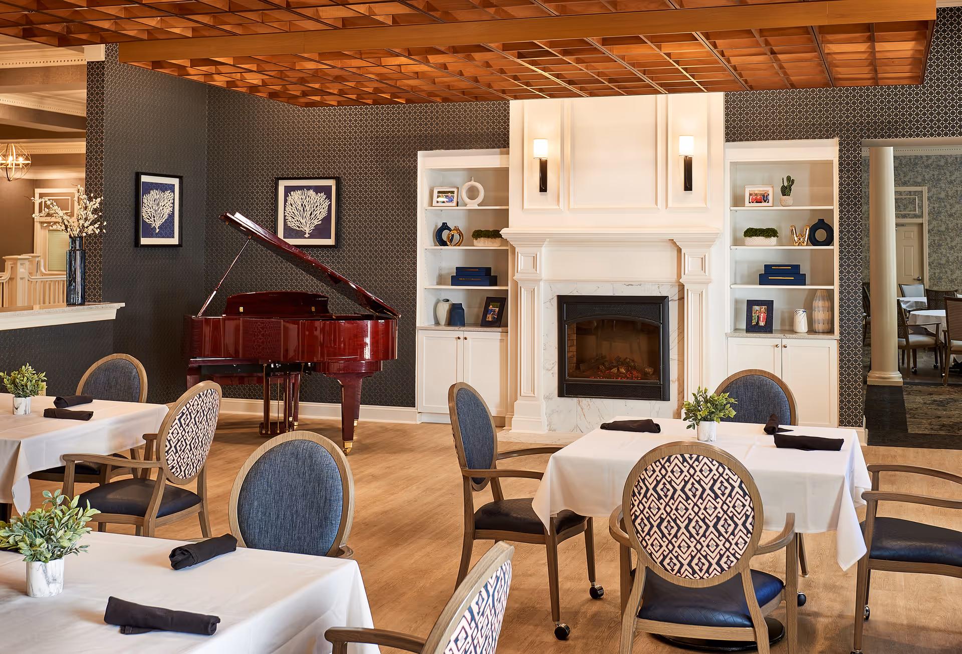 A cozy dining area with tables covered in white tablecloths and black napkins, surrounded by cushioned chairs with patterned backs. A red grand piano is positioned against a dark patterned wall with two framed coral artworks above it. In the center, there is a white fireplace with built-in shelves on either side, decorated with books, plants, and small decorative items. The room has a wooden floor and a ceiling with a wooden grid design.