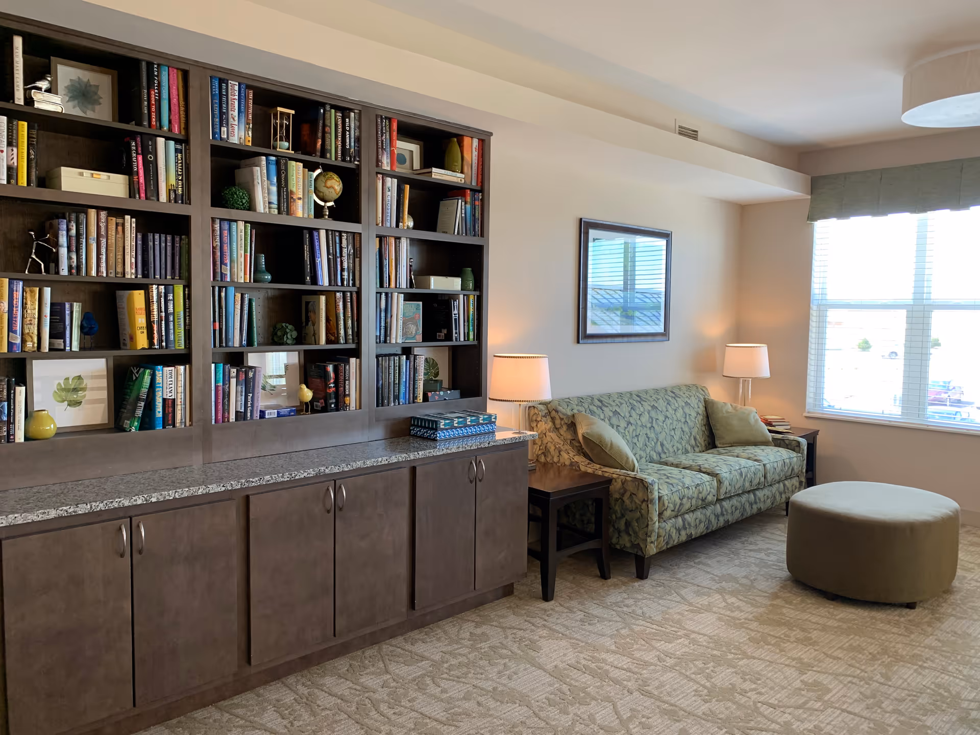 A cozy living room area with a large built-in bookshelf filled with books and decorative items on the left. To the right, there is a patterned green sofa with two matching pillows, a round green ottoman, two side tables each with a lamp, and a window with blinds letting in natural light.