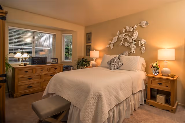 A cozy bedroom with a neatly made bed covered in a white quilt and decorative pillows. On either side of the bed are wooden nightstands with lamps, a clock, and a small flower arrangement. Above the bed is a decorative wall art piece featuring large white leaves. A wooden dresser with multiple drawers is positioned under a window with blinds, and a small cushioned bench is placed at the foot of the bed. The room is warmly lit and has a comfortable, inviting atmosphere.