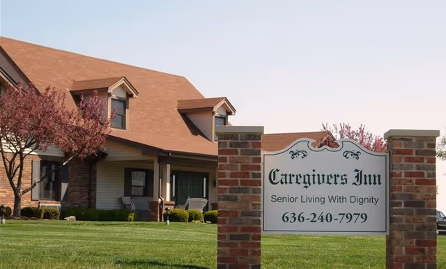 Exterior view of Caregivers Inn, a senior living facility with a brick and siding building, a well-maintained lawn, and blossoming trees. A sign in the foreground displays the facility name, slogan, and phone number.