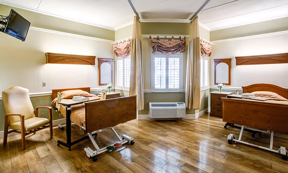 Sunlit shared senior care bedroom with two adjustable hospital beds, a reclining chair, wooden floors, and a central window with curtains.