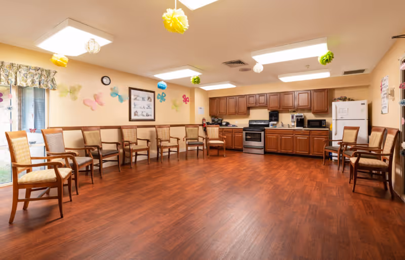 A spacious room with wooden flooring and beige walls decorated with colorful paper butterflies and hanging pom-poms. The room features a row of wooden chairs with cushioned seats arranged along the walls. At the far end, there is a kitchenette area with wooden cabinets, a stove, a refrigerator, and various kitchen appliances. A window with floral curtains is visible on the left side.