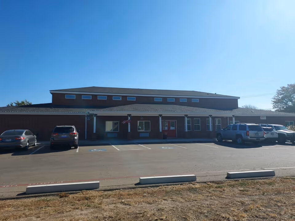 Front view of a single-story brick senior living facility with an American flag by the entrance and cars parked in the lot.