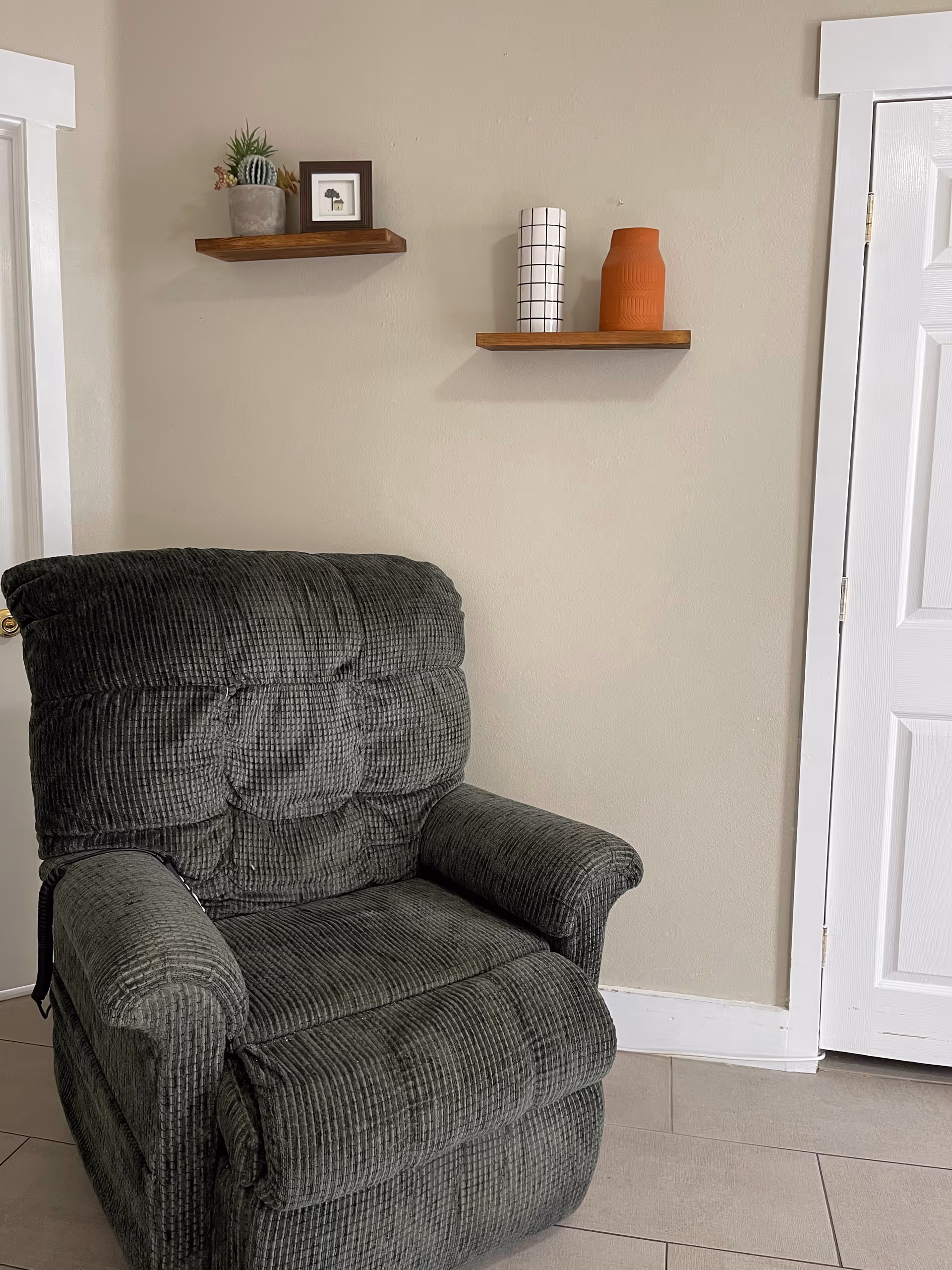 A dark green cushioned recliner chair positioned against a beige wall with two small wooden floating shelves mounted above it. The shelves hold decorative items including a small potted cactus, a framed picture, a white vase with a black grid pattern, and an orange vase. A white door is partially visible on the right side of the image.