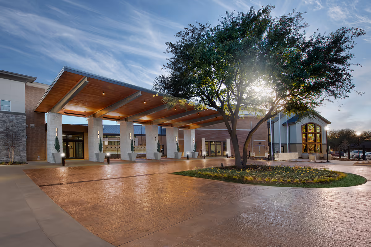 Exterior view of Presbyterian Village North showing the entrance with a large covered drop-off area supported by stone pillars, a tree with sunlight shining through its branches, and a landscaped circular garden bed in front.