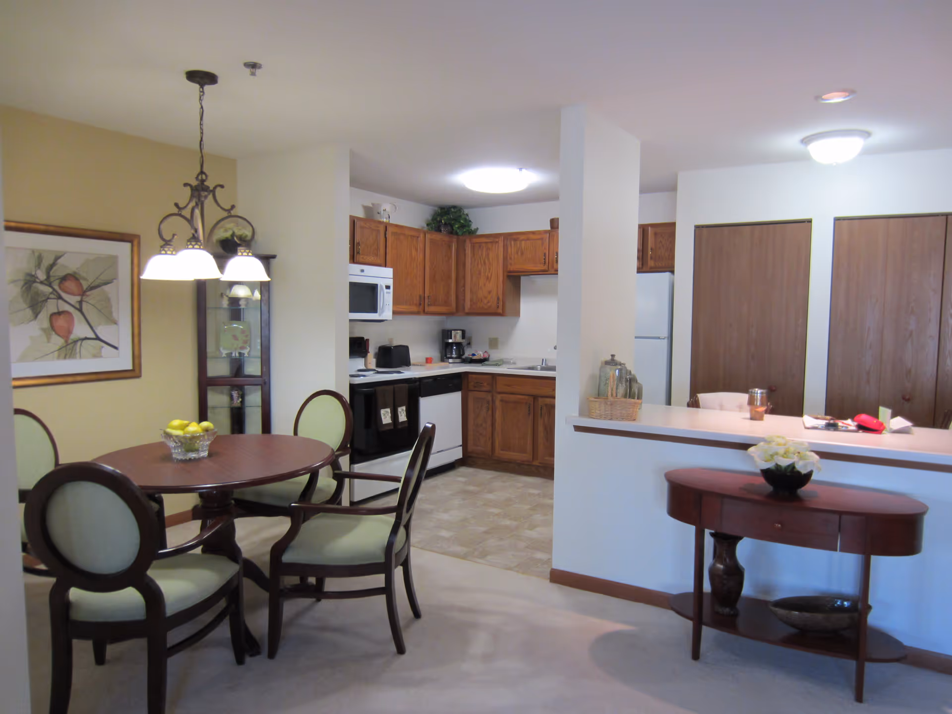 Open-plan interior with a round dining table and chairs in the foreground and a kitchen with wooden cabinets and appliances in the background.