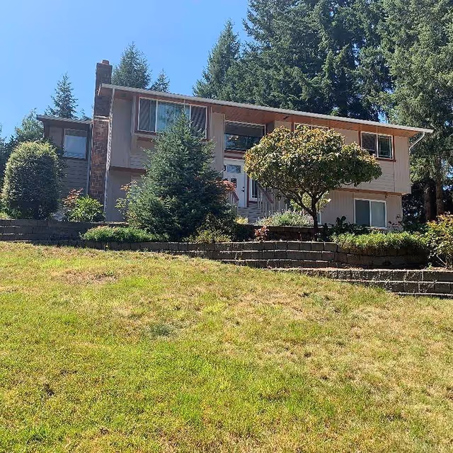 Front exterior of a two-story house with a sloped lawn, terraced stone planters, and trees.