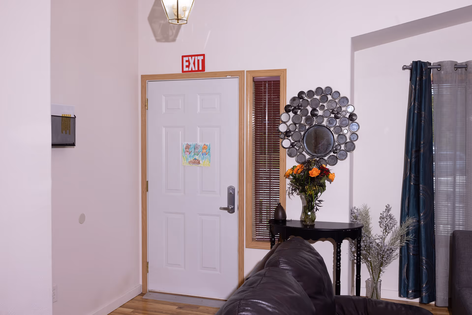 Interior view of a living room area showing a white exit door with a child's drawing taped to it. To the right of the door is a small black table with a vase of orange flowers and decorative items. Above the table is a circular wall mirror with multiple smaller round mirrors around it. A dark leather couch is partially visible in the foreground, and blue curtains hang on the window to the right.