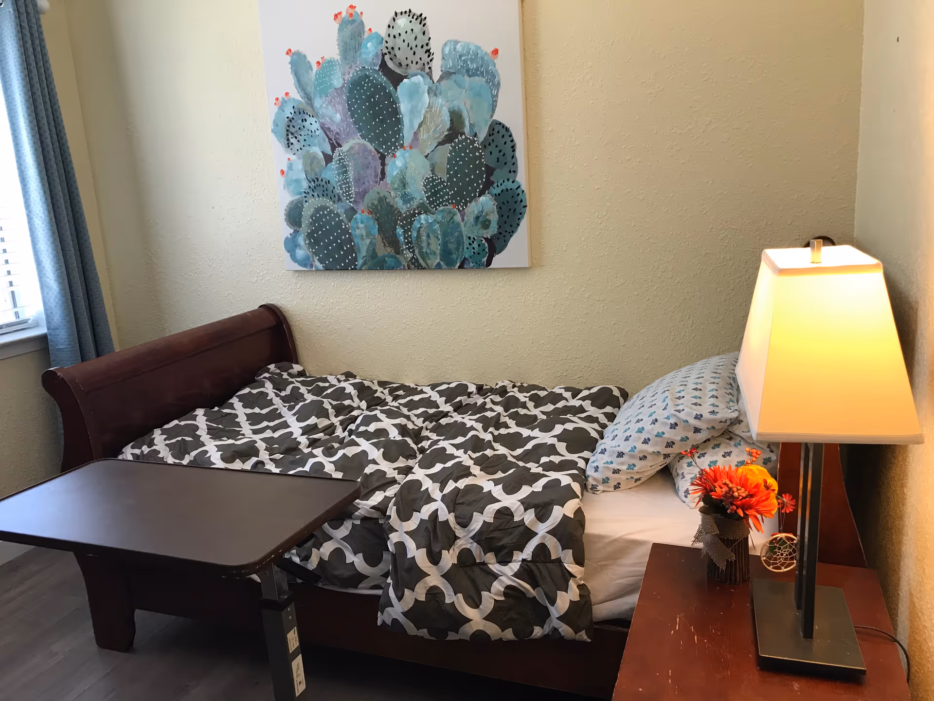 A small bedroom with a wooden bed featuring a patterned gray and white comforter and a pillow with a blue design. A wooden bedside table holds a lamp with a rectangular shade and a small vase with orange flowers. A colorful cactus painting hangs on the beige wall above the bed. A window with blue curtains is partially visible on the left side.