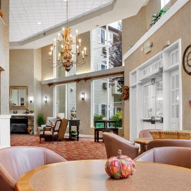 Bright, furnished senior living lobby with round tables and chairs in the foreground, a chandelier, oversized windows, and a seating area near a fireplace.