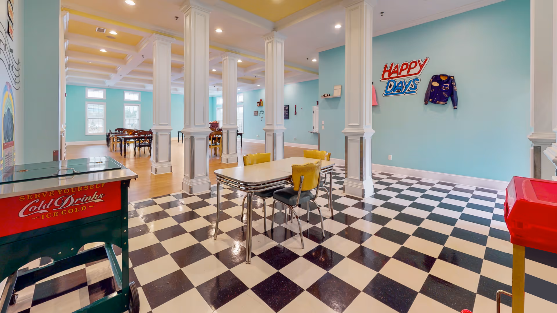 Bright common dining/activity room with teal walls, black-and-white checkered floor, retro diner tables and chairs, white columns and a 'Happy Days' sign on the wall.