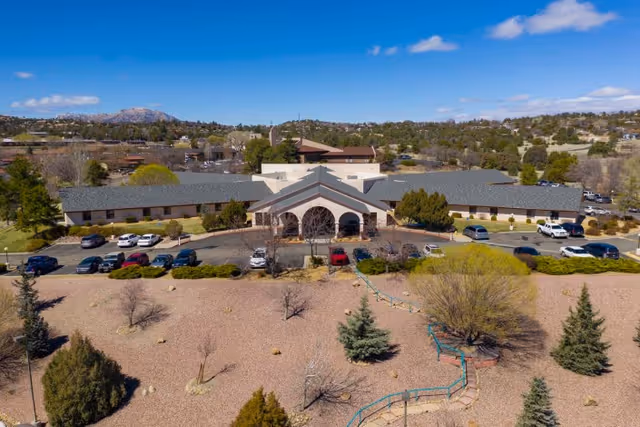 Aerial view of a single-story senior living facility with an arched main entrance, parking lot, and landscaped front grounds under a blue sky.