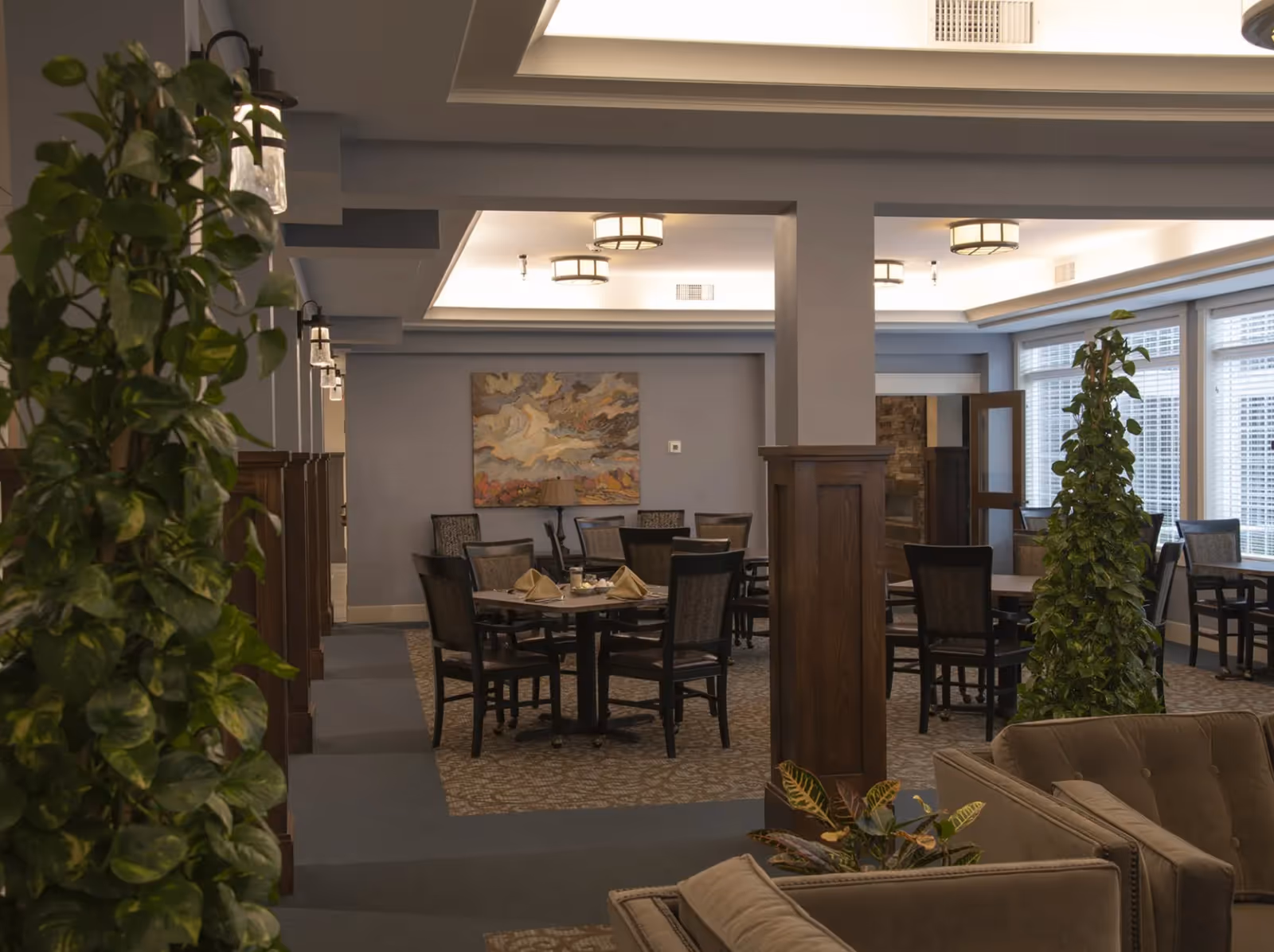 Communal dining area with round tables and chairs, upholstered seating, potted plants, and ceiling lights.