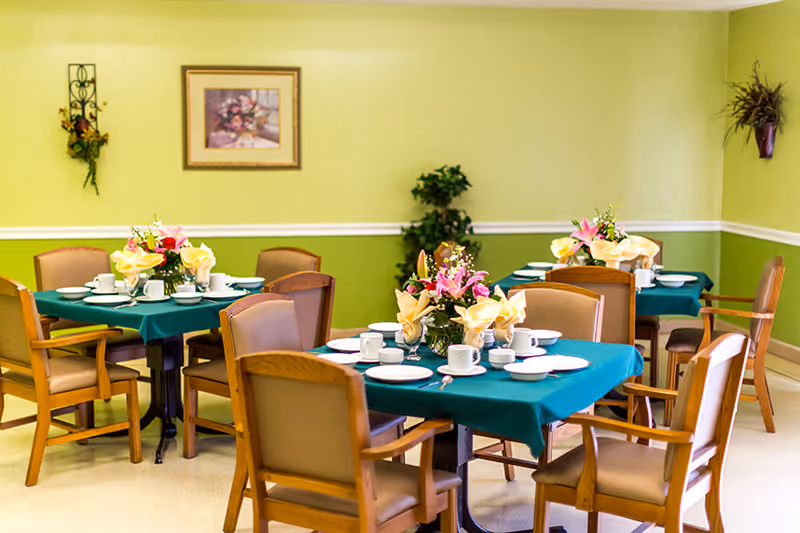 A dining room with three tables covered in teal tablecloths, each set with white plates, cups, and silverware. Each table has a floral centerpiece with pink and yellow flowers. The room has light green walls with white trim, a framed floral painting, and some wall-mounted plants and greenery in the corners.