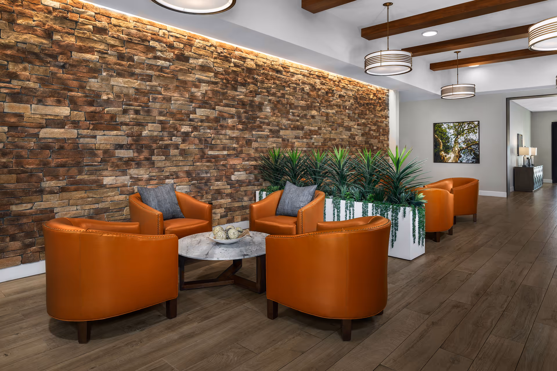 A modern lounge with orange leather armchairs arranged around a round marble table in front of a stone accent wall and planter divider.