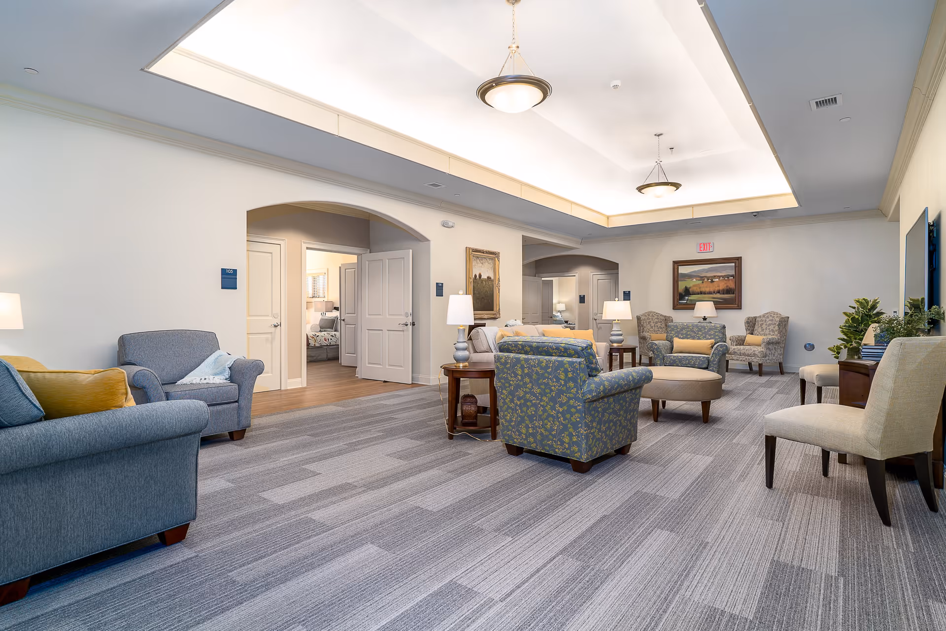 A spacious and well-lit common living area in a senior living facility with multiple upholstered armchairs and sofas arranged around small tables. The room features a patterned carpet, soft lighting from ceiling fixtures, framed artwork on the walls, and an open doorway revealing a bedroom in the background.