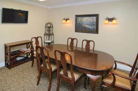 A dining room with an oval wooden table, eight chairs, wall art, and a wall-mounted TV.