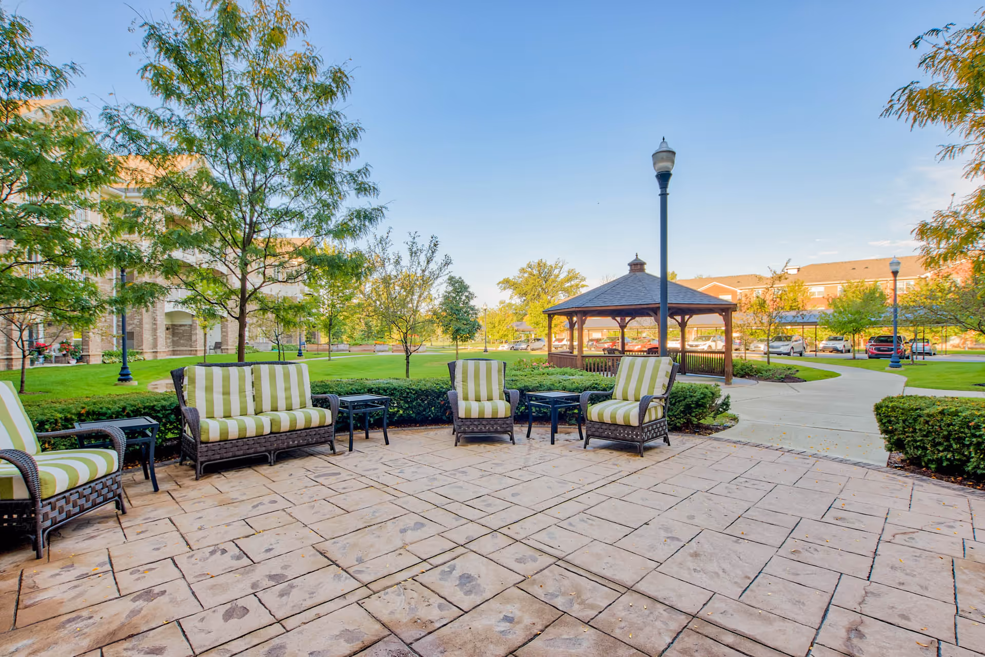 Outdoor patio area at Rose Senior Living Clinton Township with cushioned wicker chairs and small tables arranged on a stone-paved surface, surrounded by green grass, trees, and a wooden gazebo in the background under a clear blue sky.