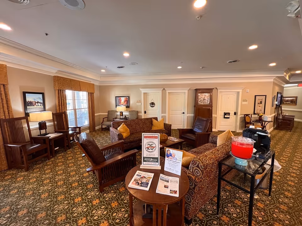 A cozy senior living facility common area with patterned carpet, multiple armchairs and sofas with yellow cushions, wooden chairs, and side tables. There are framed pictures on the walls, windows with curtains, and a small table with informational brochures and a sign celebrating 25 years. A drink dispenser with red liquid and a coffee maker are on a small black cart to the right.