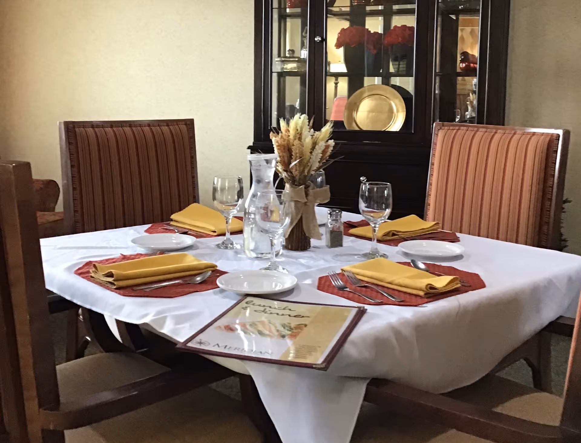 A dining table set for four with a white tablecloth, red placemats, yellow folded napkins, plates, forks, spoons, and wine glasses. A glass pitcher of water and a centerpiece with dried flowers are on the table. In the background, there is a dark wooden china cabinet with decorative plates and red flowers inside.