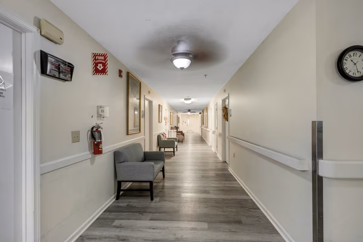 A long, well-lit hallway in a senior living facility with light-colored walls, wood-style flooring, ceiling fans with lights, and several chairs placed along the walls. There are framed pictures, a fire extinguisher, and a clock on the walls.