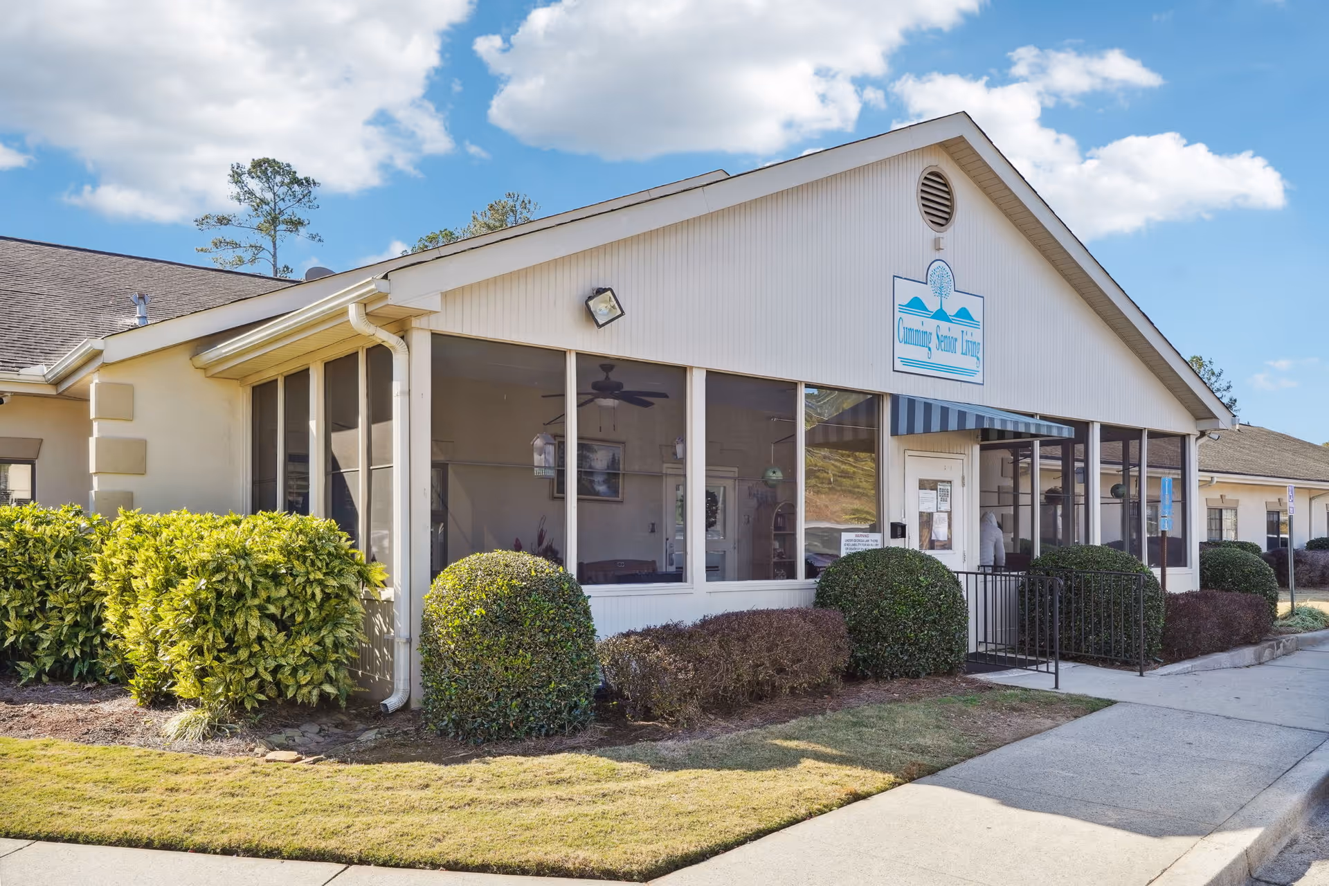 Exterior view of a single-story building with large windows and a sign that reads 'Cumming Senior Living'. The building is surrounded by neatly trimmed bushes and a well-maintained lawn under a partly cloudy sky.