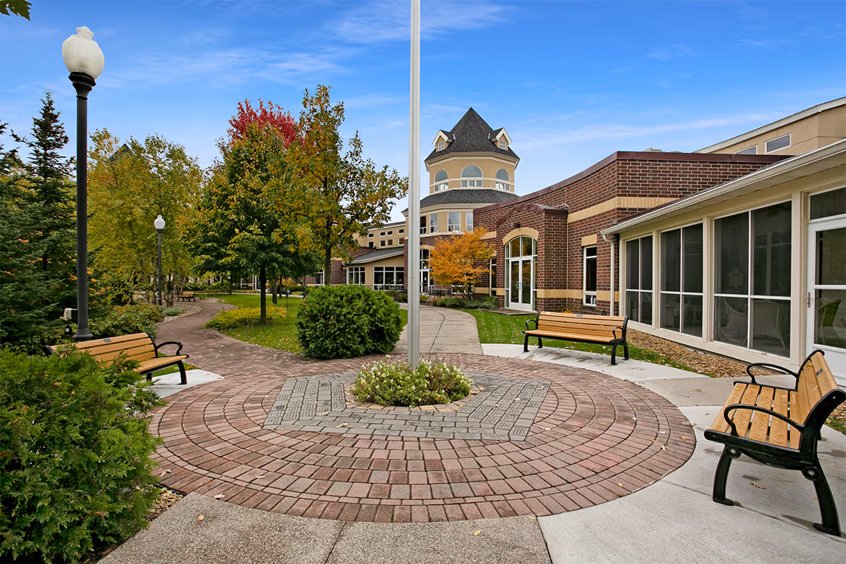 Outdoor courtyard area at Boutwells Landing featuring a circular brick-paved area with a flagpole in the center, surrounded by benches and landscaped greenery including trees with autumn foliage. The building with brick and beige exterior walls is visible in the background under a clear blue sky.