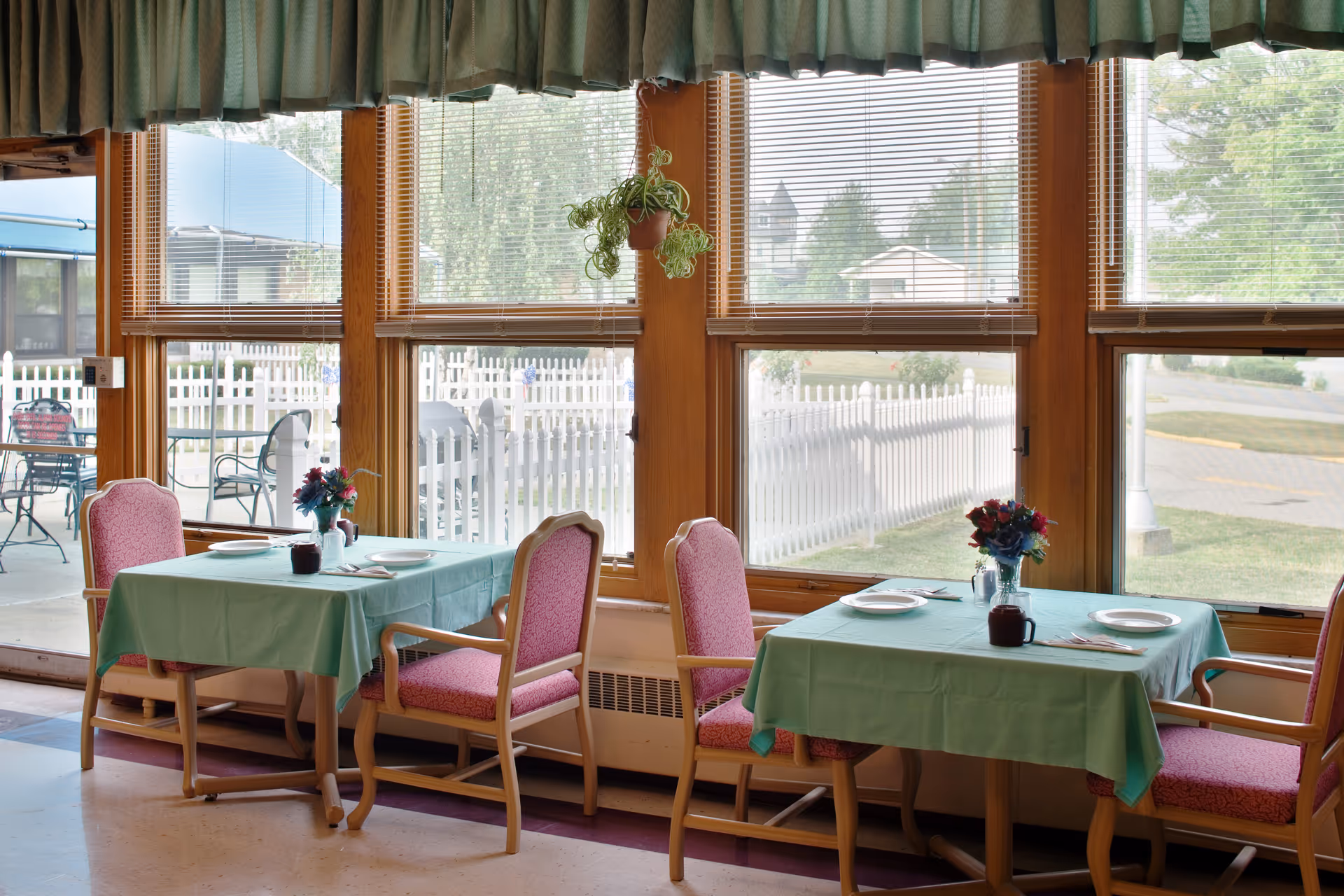 Two small dining tables with green tablecloths and pink chairs set by large windows overlooking a white picket fence.