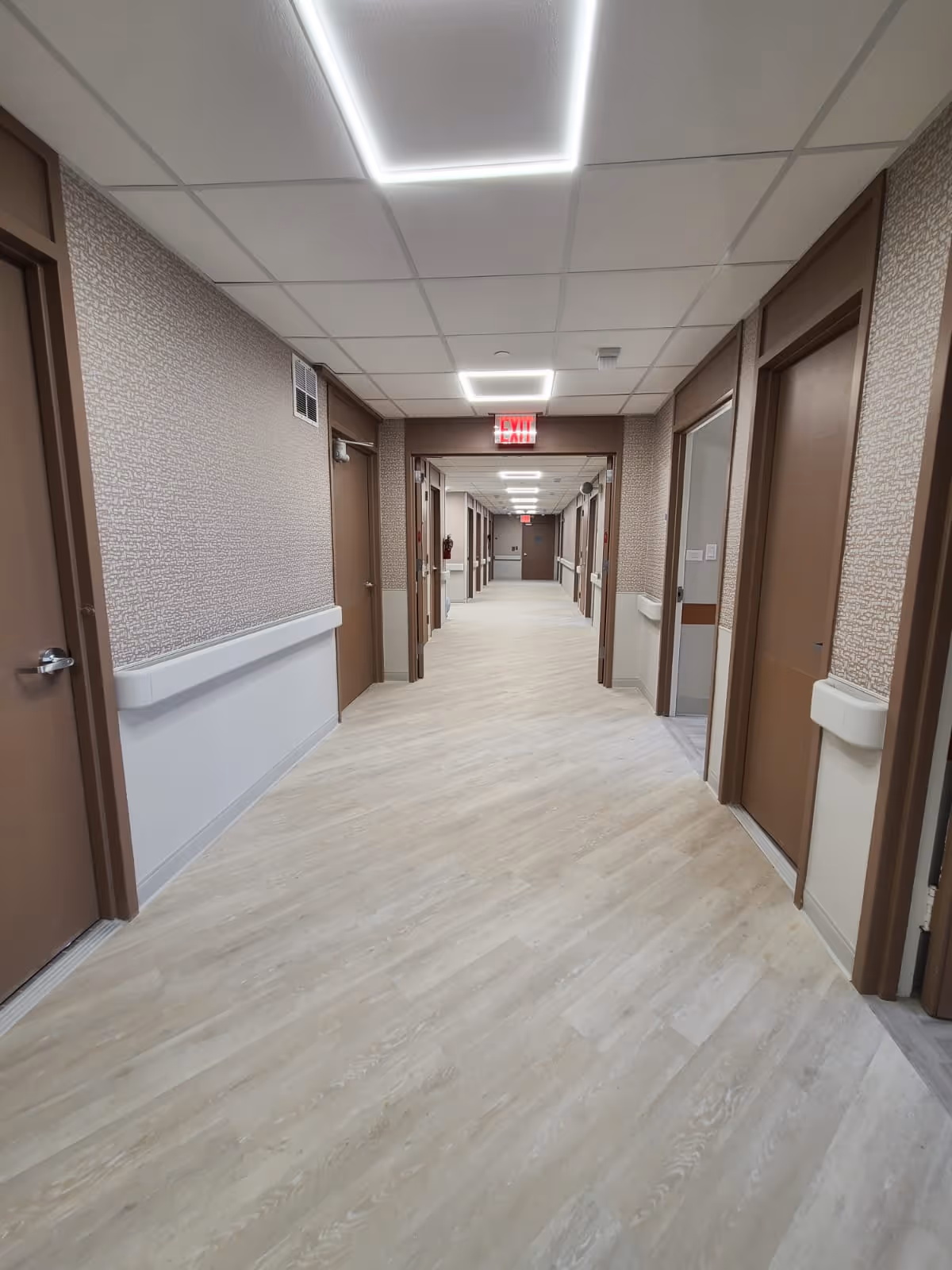 A long, well-lit hallway in a healthcare or senior living facility with multiple closed brown doors on both sides. The floor has light wood-like vinyl flooring, and the walls are decorated with textured wallpaper and white handrails. Ceiling lights are rectangular and bright, and an illuminated red exit sign is visible at the far end of the corridor.