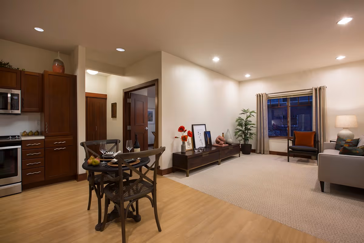 A cozy living area and kitchen space in a senior living facility. The kitchen features wooden cabinets, a microwave, and a stove. A small round dining table with two chairs is set with wine glasses, a bottle of wine, and pears. The living room has a beige carpet, a wooden console table with decorative items and framed pictures, a potted plant, a window with beige curtains, a cushioned armchair with an orange pillow, and a gray sofa with colorful pillows. The room is softly lit with recessed ceiling lights.
