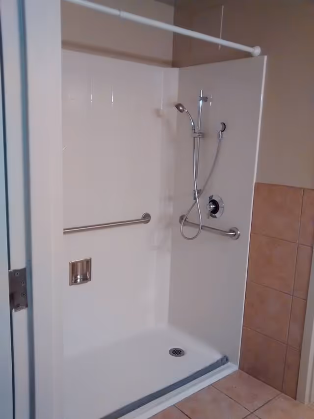 Walk-in shower stall with a handheld showerhead, horizontal grab bars, and a tiled floor.