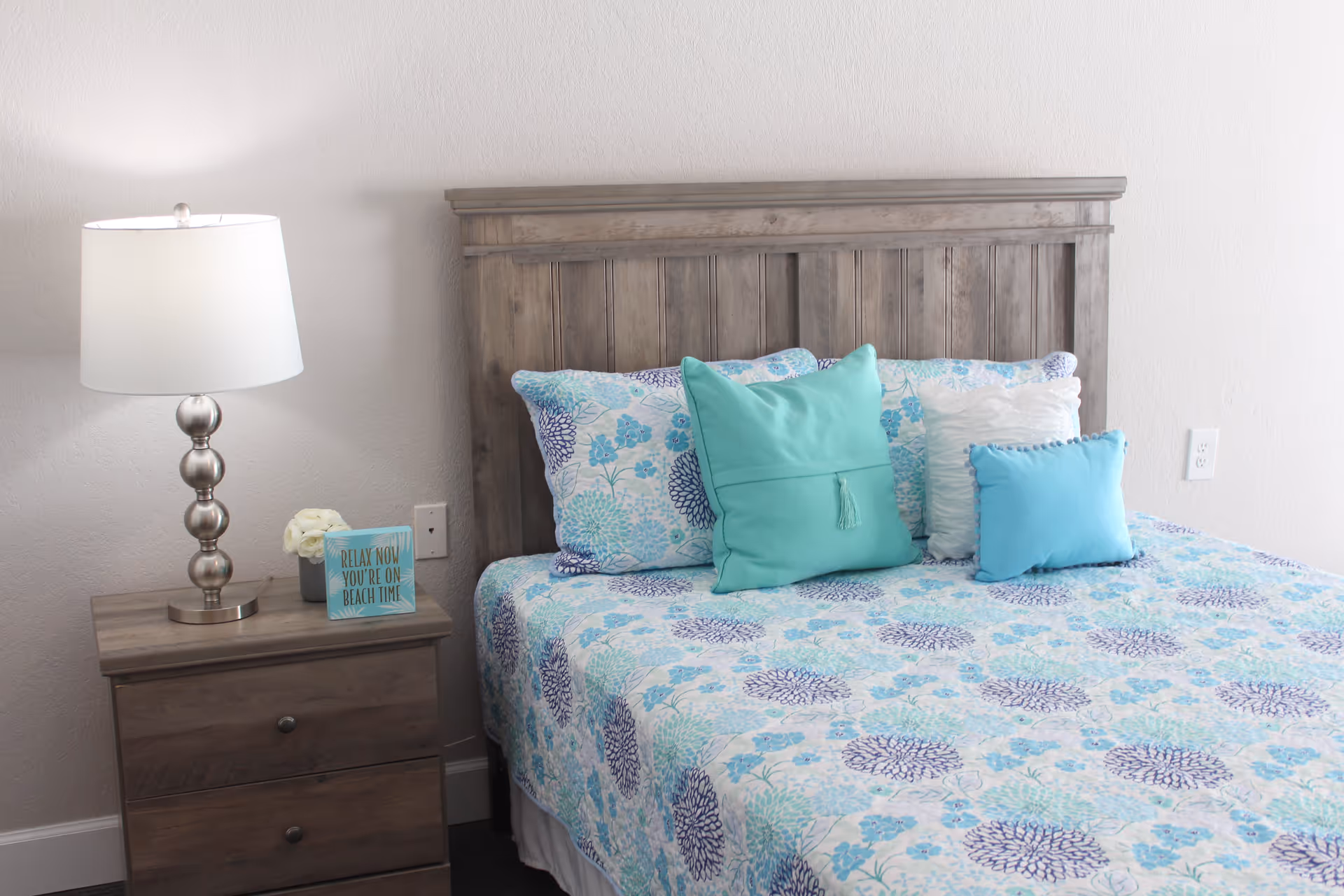 A bedroom with a wooden headboard bed dressed in a blue and white floral patterned bedspread and multiple pillows in shades of blue and white. Next to the bed is a wooden nightstand with two drawers, a silver lamp with a white shade, a small flower arrangement, and a decorative sign that reads 'RELAX NOW YOU'RE ON BEACH TIME'.