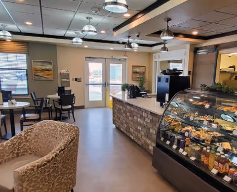 Interior view of a dining area with several tables and chairs, a comfortable armchair in the foreground, a counter with a stone facade, and a glass display case filled with various food items and beverages. The room has pendant lights hanging from the ceiling, framed artwork on the walls, and double glass doors leading outside.