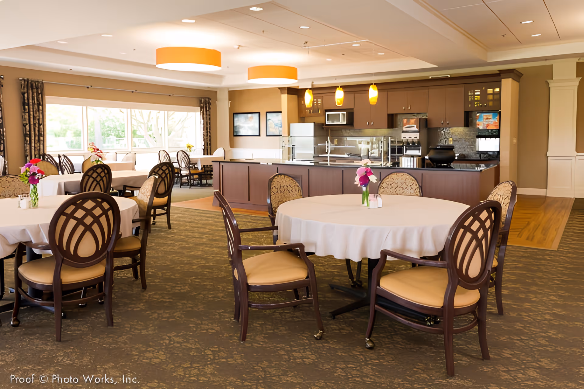 A spacious dining room with round tables covered in white tablecloths and surrounded by wooden chairs with cushioned seats and decorative backs. The room features large windows with curtains allowing natural light to fill the space. In the background, there is a kitchen area with brown cabinetry, stainless steel appliances, and pendant lights hanging from the ceiling. The carpeted floor has a patterned design, and there are small flower arrangements on the tables.