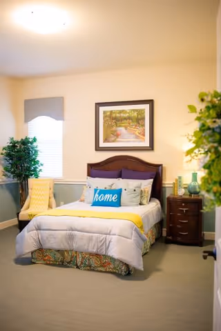 A cozy bedroom with a neatly made bed featuring a 'home' pillow, a yellow throw blanket, and multiple pillows. There is a wooden headboard, a framed picture of a garden pathway on the wall above the bed, a wooden nightstand with decorative items, a yellow armchair with a yellow blanket, and a potted plant near the window with a gray valance.