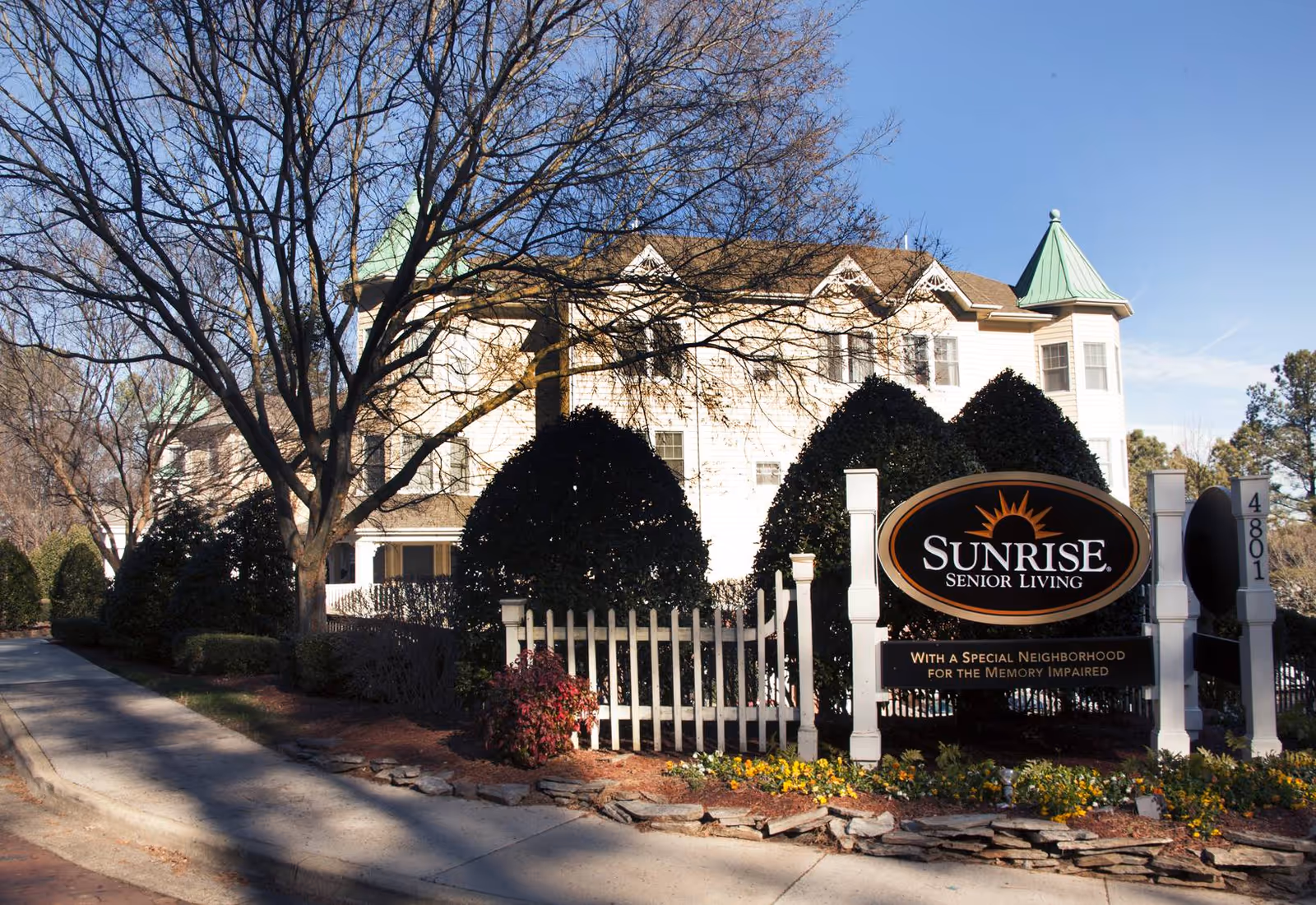 Front view of the Sunrise Senior Living building and entrance sign framed by a white picket fence, shrubs and a driveway.