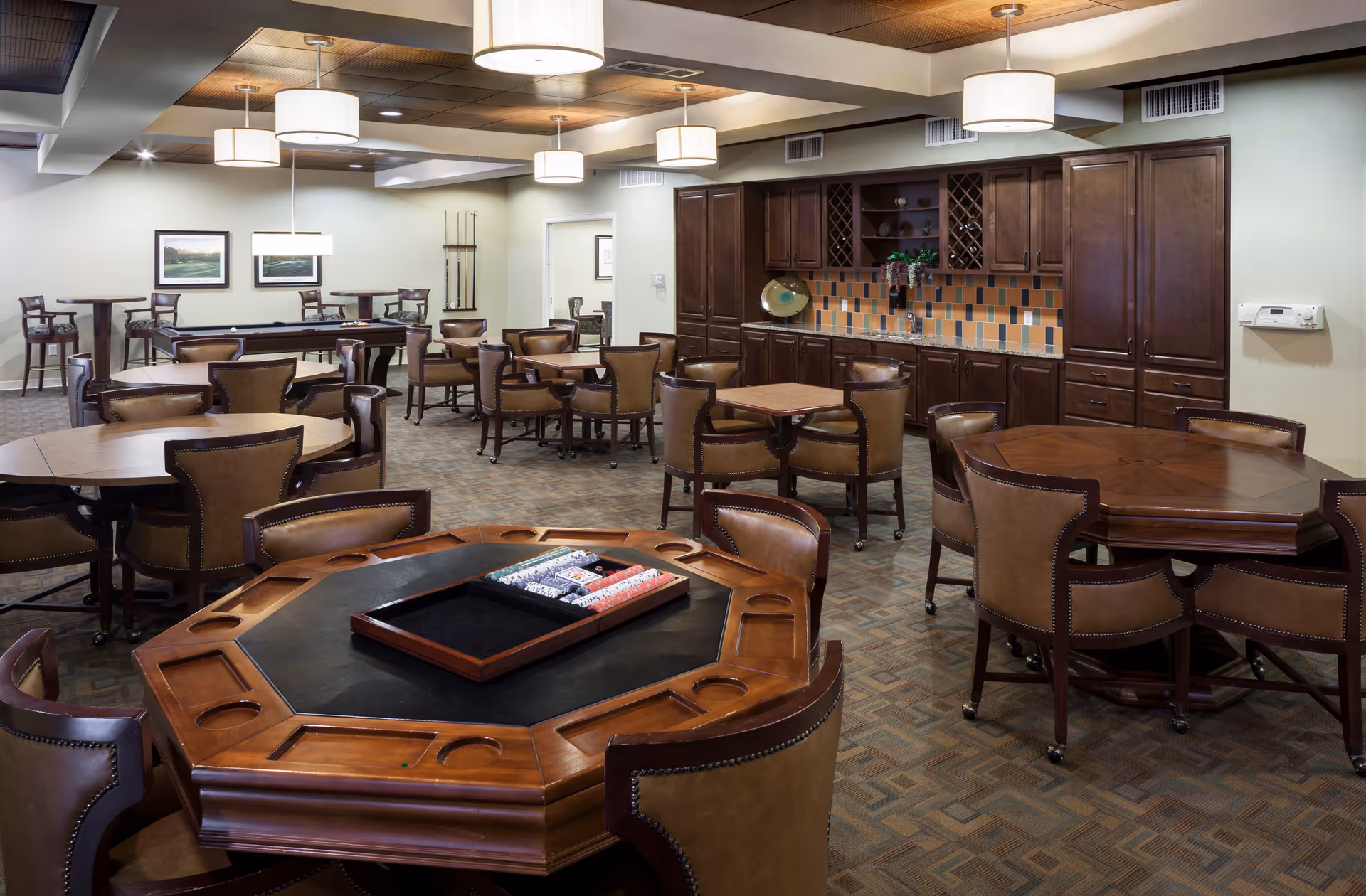 A spacious common room with multiple wooden tables and leather chairs arranged for group activities and dining. The foreground features a large octagonal game table with poker chips and card trays. The background shows a pool table, additional seating, and a kitchenette area with dark wood cabinets and a tiled backsplash. The room is well-lit with ceiling lights and has a warm, inviting atmosphere.