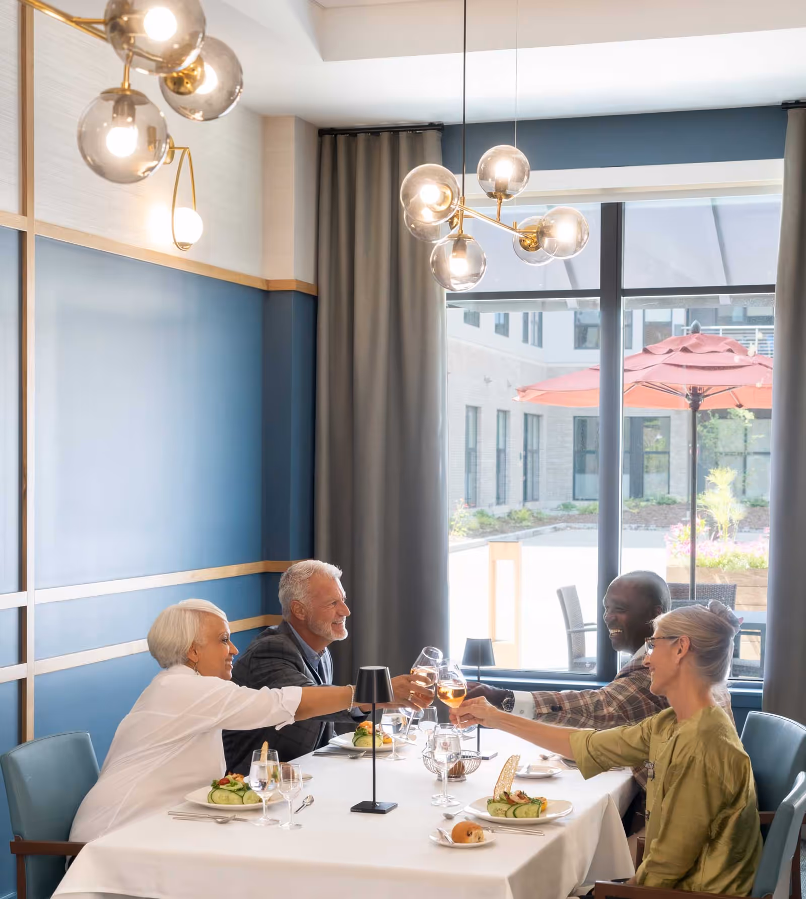 Four older adults seated at a table in a bright dining room clinking wine glasses.