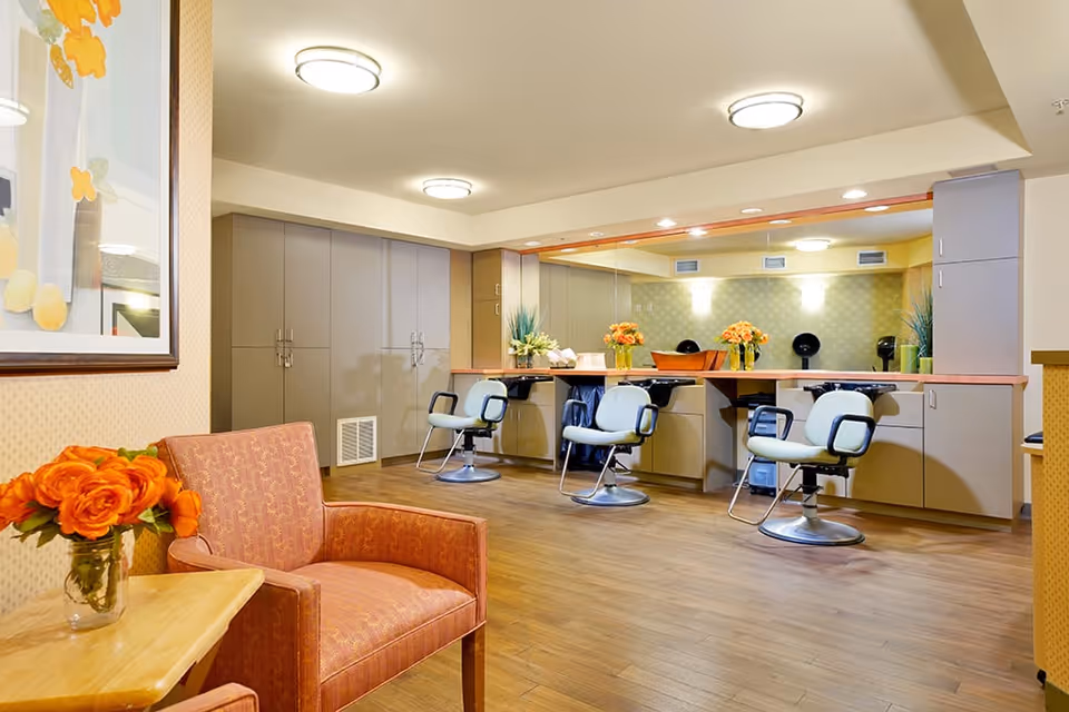 Interior view of a salon area in a senior living facility featuring three salon chairs in front of a large mirror with a countertop. There are cabinets and drawers beneath the countertop and a wooden floor. In the foreground, there is an orange upholstered armchair next to a small wooden table with a vase of orange flowers. The walls are light-colored with soft lighting from ceiling fixtures.