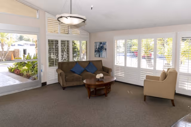 Sunlit common living area with a sofa, armchair and round coffee table in front of large shuttered windows overlooking outdoor landscaping.